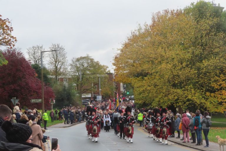 Pipers head a procession along the road, lined with lookers-on